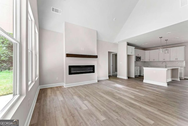 unfurnished living room featuring high vaulted ceiling, light wood-style flooring, a sink, visible vents, and a glass covered fireplace