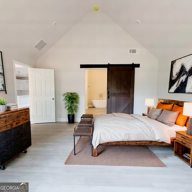 bedroom with a barn door, visible vents, ensuite bath, light wood-type flooring, and high vaulted ceiling