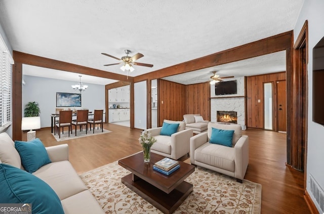 living room with hardwood / wood-style flooring, ceiling fan with notable chandelier, a textured ceiling, a stone fireplace, and wood walls