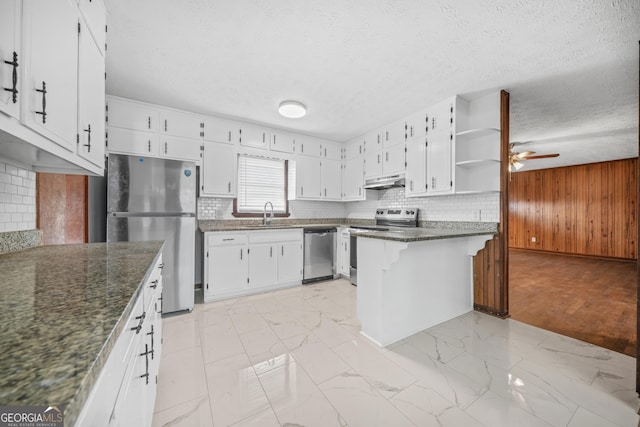 kitchen featuring ceiling fan, appliances with stainless steel finishes, dark stone countertops, wooden walls, and white cabinets