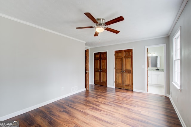 unfurnished bedroom with crown molding, ensuite bath, ceiling fan, wood-type flooring, and multiple closets