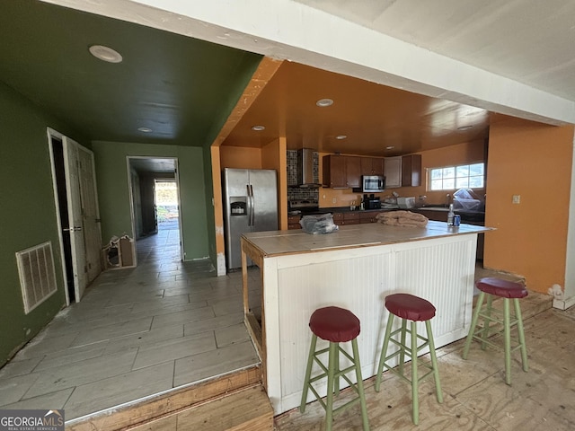 kitchen with appliances with stainless steel finishes, kitchen peninsula, a kitchen bar, and wall chimney exhaust hood