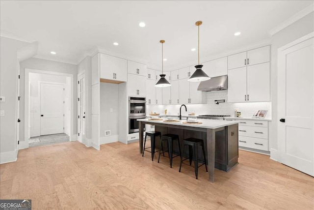 kitchen with appliances with stainless steel finishes, white cabinetry, hanging light fixtures, a kitchen island with sink, and wall chimney range hood