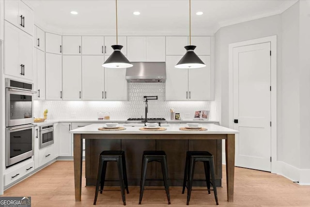 kitchen featuring wall chimney range hood, appliances with stainless steel finishes, white cabinetry, an island with sink, and decorative light fixtures