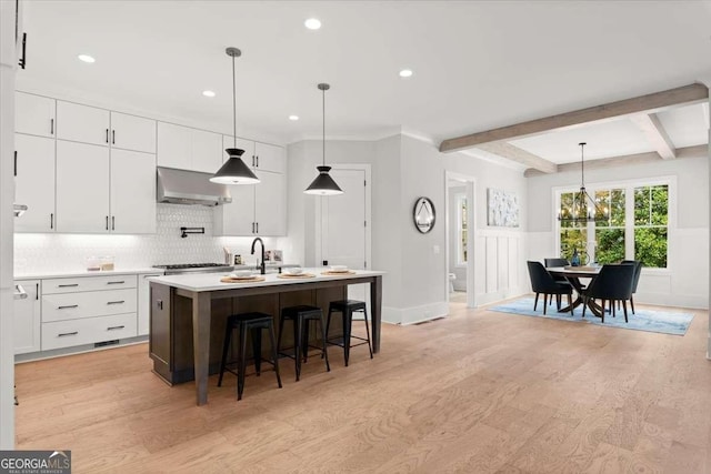 kitchen featuring hanging light fixtures, white cabinetry, a kitchen island with sink, and wall chimney range hood