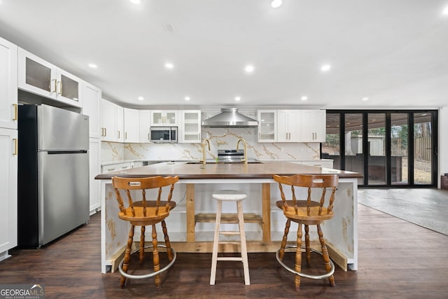 kitchen featuring white cabinetry, a center island with sink, appliances with stainless steel finishes, decorative backsplash, and wall chimney range hood