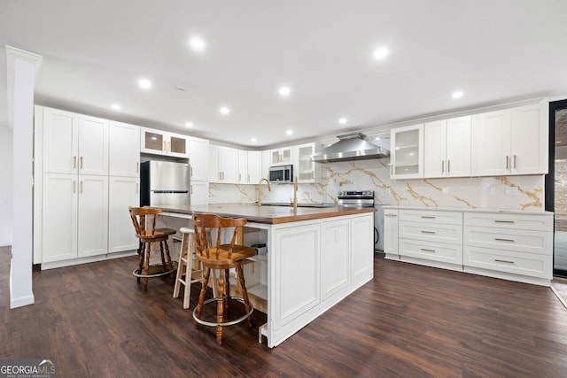 kitchen featuring a kitchen island with sink, wall chimney range hood, white cabinetry, and stainless steel appliances