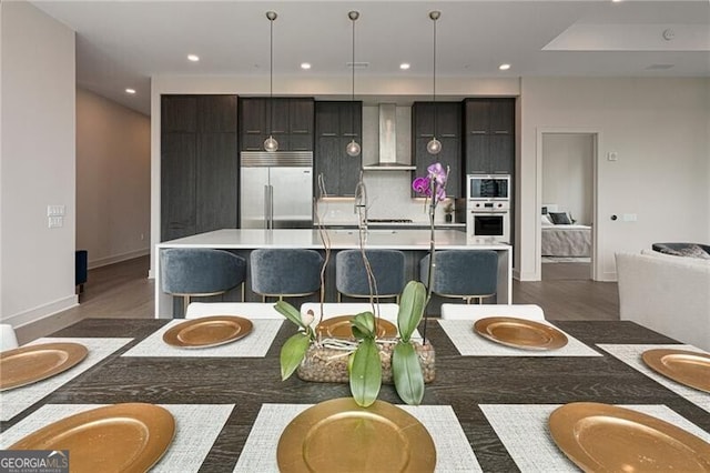 kitchen with decorative light fixtures, a center island with sink, a kitchen breakfast bar, stainless steel appliances, and wall chimney range hood