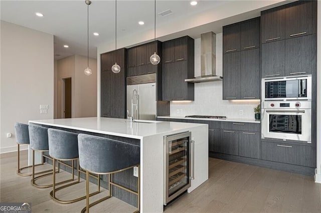 kitchen featuring hanging light fixtures, built in appliances, wine cooler, a center island with sink, and wall chimney exhaust hood