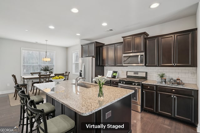 kitchen with an island with sink, sink, hanging light fixtures, stainless steel appliances, and dark brown cabinets