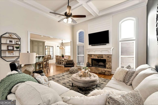 living room with a stone fireplace, ceiling fan with notable chandelier, hardwood / wood-style flooring, coffered ceiling, and crown molding