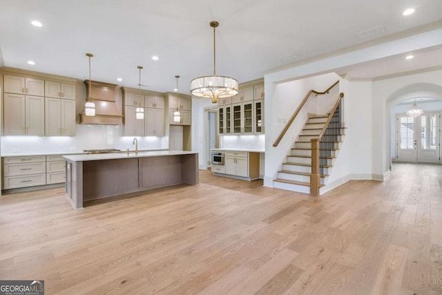 kitchen featuring pendant lighting, a chandelier, custom exhaust hood, light hardwood / wood-style floors, and a center island with sink