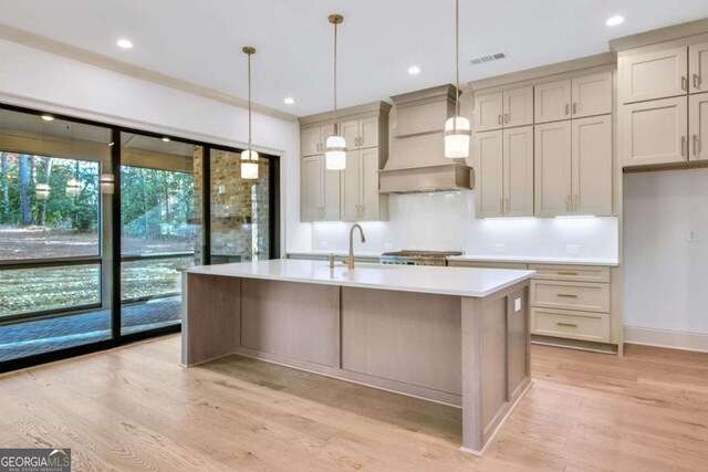 kitchen with sink, hanging light fixtures, a kitchen island with sink, custom range hood, and light hardwood / wood-style flooring