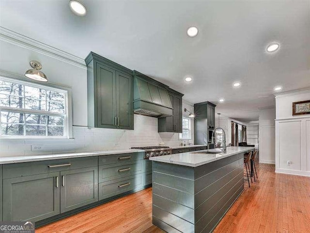 kitchen featuring custom exhaust hood, tasteful backsplash, crown molding, an island with sink, and light hardwood / wood-style floors