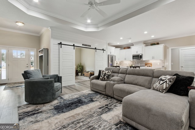 living room featuring ceiling fan, a tray ceiling, ornamental molding, light hardwood / wood-style floors, and a barn door