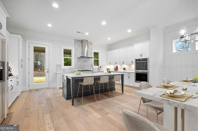 kitchen with double oven, tasteful backsplash, white cabinets, a kitchen bar, and wall chimney range hood