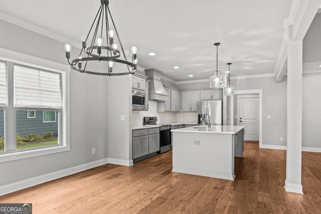 kitchen with gray cabinetry, decorative light fixtures, appliances with stainless steel finishes, custom range hood, and a kitchen island with sink