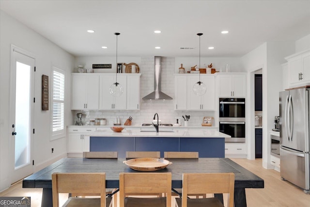 kitchen with a center island with sink, wall chimney range hood, stainless steel refrigerator, and decorative light fixtures