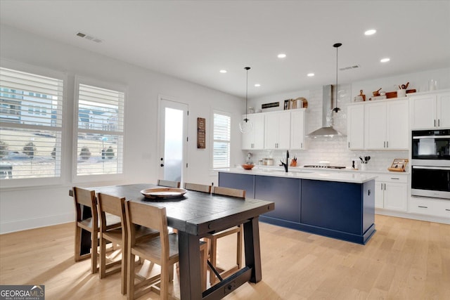 kitchen featuring double oven, decorative light fixtures, an island with sink, white cabinets, and wall chimney exhaust hood