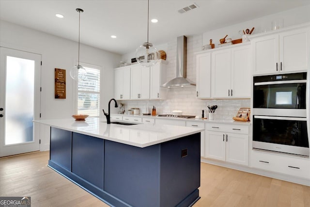 kitchen featuring wall chimney exhaust hood, sink, multiple ovens, decorative light fixtures, and an island with sink