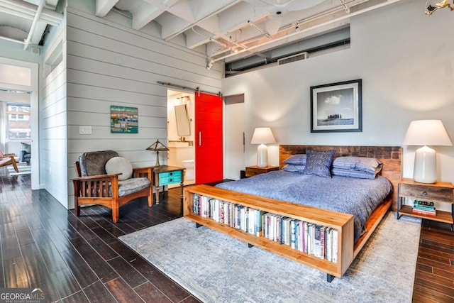 bedroom with dark wood-type flooring, connected bathroom, wooden walls, beamed ceiling, and a barn door