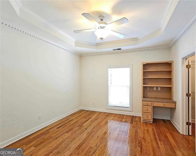 unfurnished bedroom with crown molding, wood-type flooring, built in desk, a tray ceiling, and ceiling fan