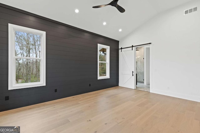 unfurnished bedroom with wooden walls, lofted ceiling, ceiling fan, a barn door, and light hardwood / wood-style flooring