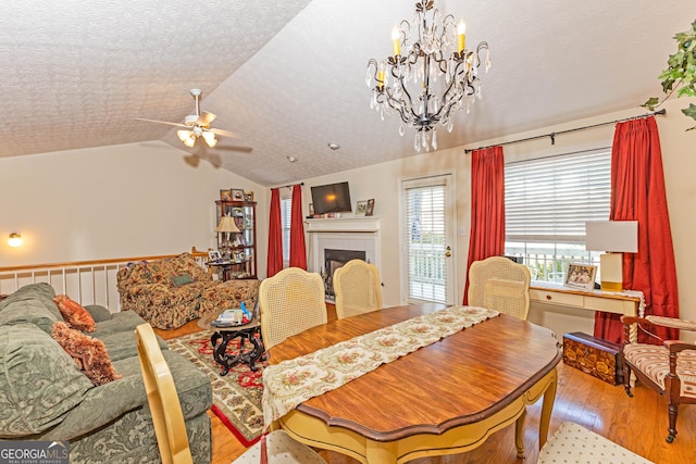 dining space featuring lofted ceiling, a textured ceiling, a tiled fireplace, ceiling fan with notable chandelier, and light wood-type flooring