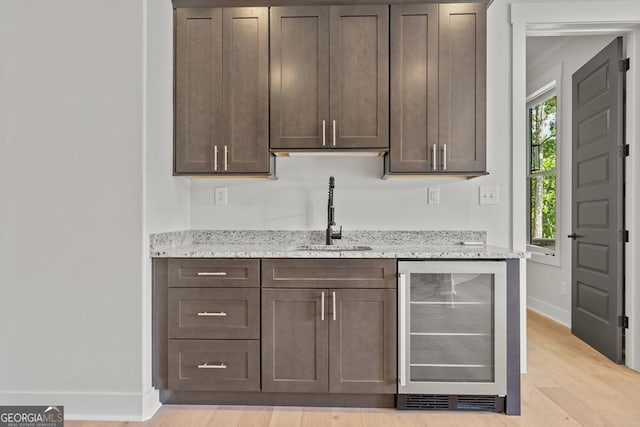 bar featuring dark brown cabinetry, sink, light stone counters, beverage cooler, and light hardwood / wood-style floors