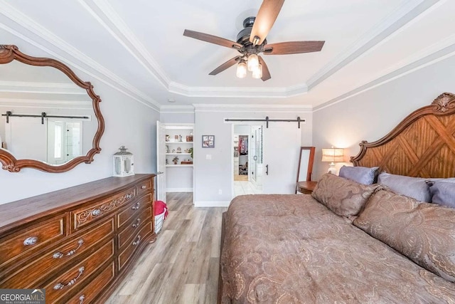 bedroom with crown molding, light hardwood / wood-style flooring, ceiling fan, a tray ceiling, and a barn door