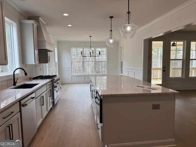 kitchen with a sink, custom exhaust hood, an inviting chandelier, a decorative wall, and appliances with stainless steel finishes