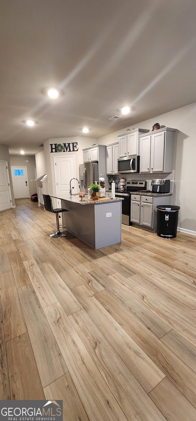 kitchen featuring appliances with stainless steel finishes, an island with sink, a kitchen breakfast bar, dark stone counters, and light hardwood / wood-style flooring