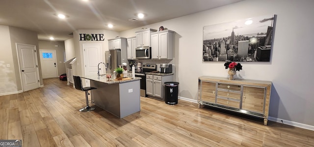 kitchen with stainless steel appliances, gray cabinets, a kitchen island with sink, and light stone countertops