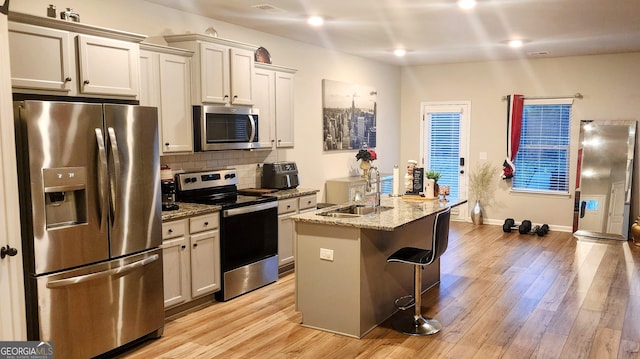 kitchen featuring sink, backsplash, stainless steel appliances, light stone counters, and an island with sink