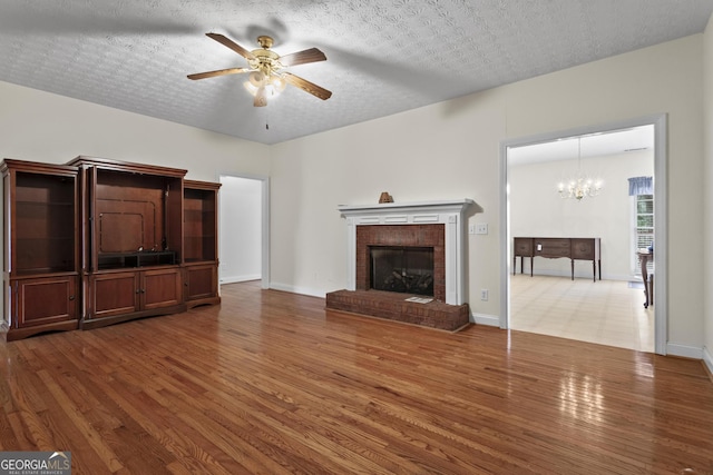 unfurnished living room with a brick fireplace, baseboards, ceiling fan with notable chandelier, wood finished floors, and a textured ceiling