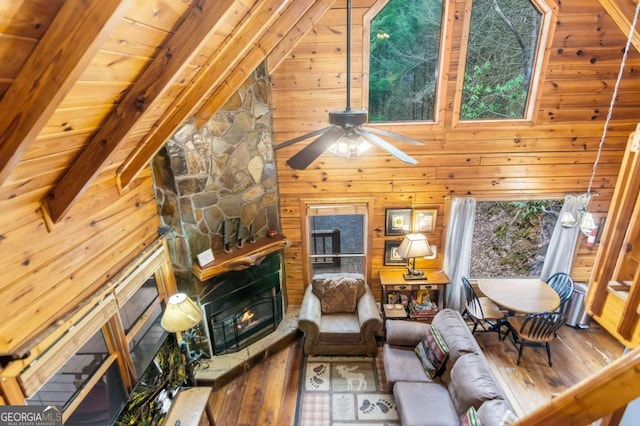 living room featuring ceiling fan, wooden walls, high vaulted ceiling, a stone fireplace, and beamed ceiling