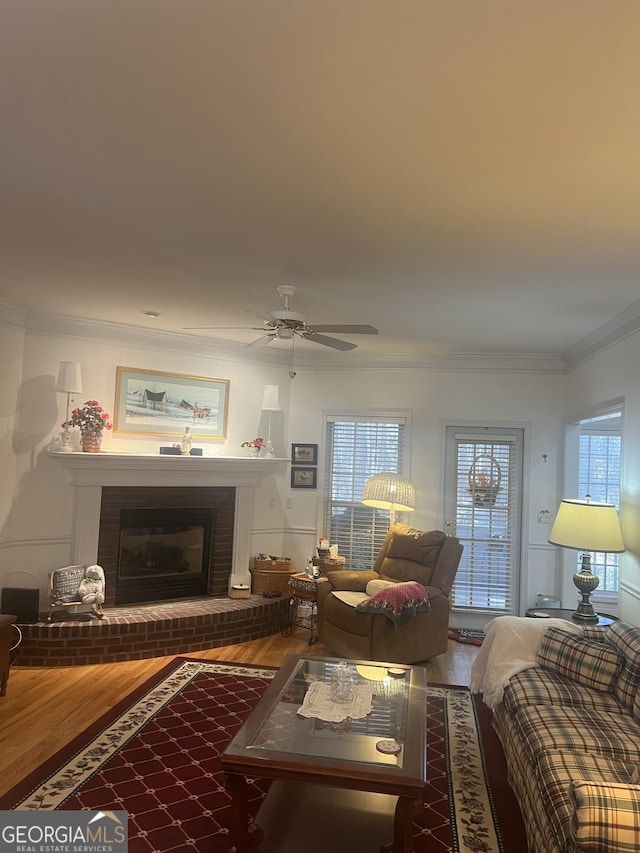 living room with ceiling fan, wood finished floors, a brick fireplace, and ornamental molding