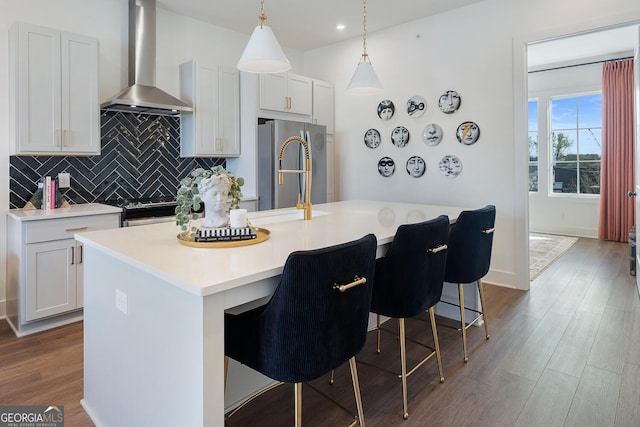 kitchen with a breakfast bar area, decorative light fixtures, a center island with sink, stainless steel appliances, and wall chimney range hood