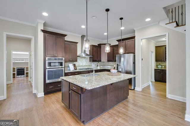 kitchen with light stone counters, appliances with stainless steel finishes, an island with sink, pendant lighting, and wall chimney range hood