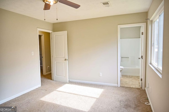 unfurnished bedroom with multiple windows, ensuite bathroom, light carpet, and ceiling fan