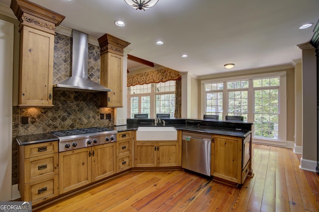 kitchen with sink, appliances with stainless steel finishes, kitchen peninsula, decorative columns, and wall chimney range hood