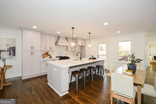 kitchen with custom exhaust hood, white cabinetry, hanging light fixtures, and a center island with sink