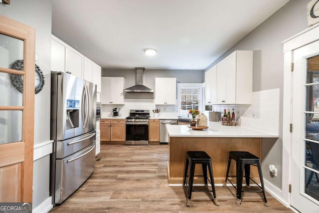 kitchen featuring a breakfast bar area, wall chimney exhaust hood, stainless steel appliances, white cabinetry, and kitchen peninsula