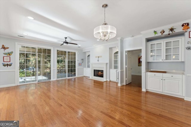 unfurnished living room with crown molding, ceiling fan with notable chandelier, built in shelves, and light hardwood / wood-style floors