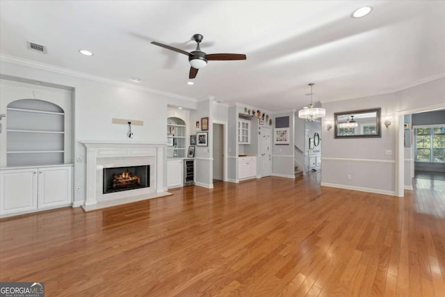 unfurnished living room featuring ceiling fan with notable chandelier, built in features, wine cooler, crown molding, and light hardwood / wood-style flooring