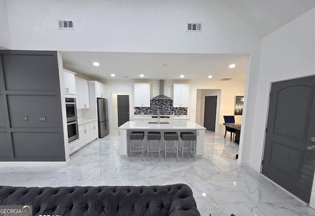 kitchen featuring a breakfast bar, stainless steel appliances, white cabinets, a center island with sink, and wall chimney exhaust hood