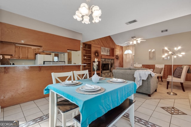 tiled dining area with vaulted ceiling, a stone fireplace, ceiling fan with notable chandelier, and wooden walls