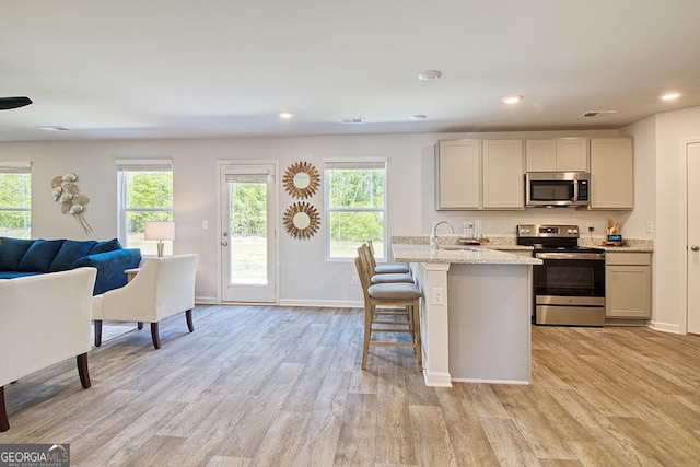 kitchen with sink, stainless steel appliances, light stone countertops, a center island with sink, and light hardwood / wood-style flooring