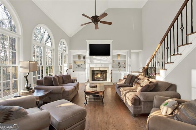 living area featuring a warm lit fireplace, a ceiling fan, wood finished floors, stairs, and high vaulted ceiling