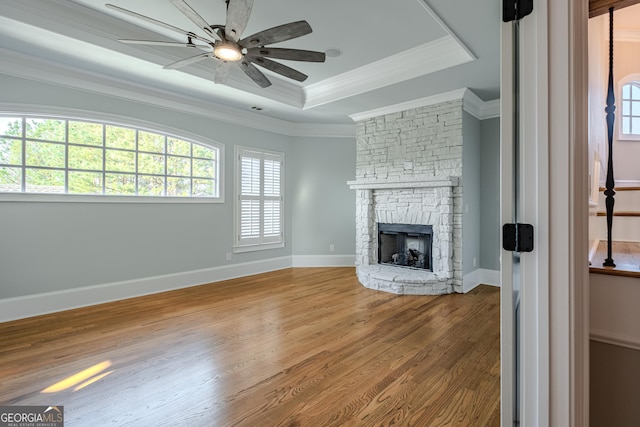 unfurnished living room with a stone fireplace, hardwood / wood-style floors, ceiling fan, a tray ceiling, and crown molding